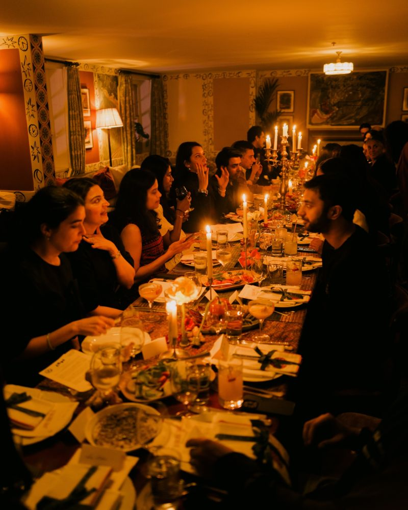 Guests in conversation along a long candlelit dinner table at Is This Seat Taken?
